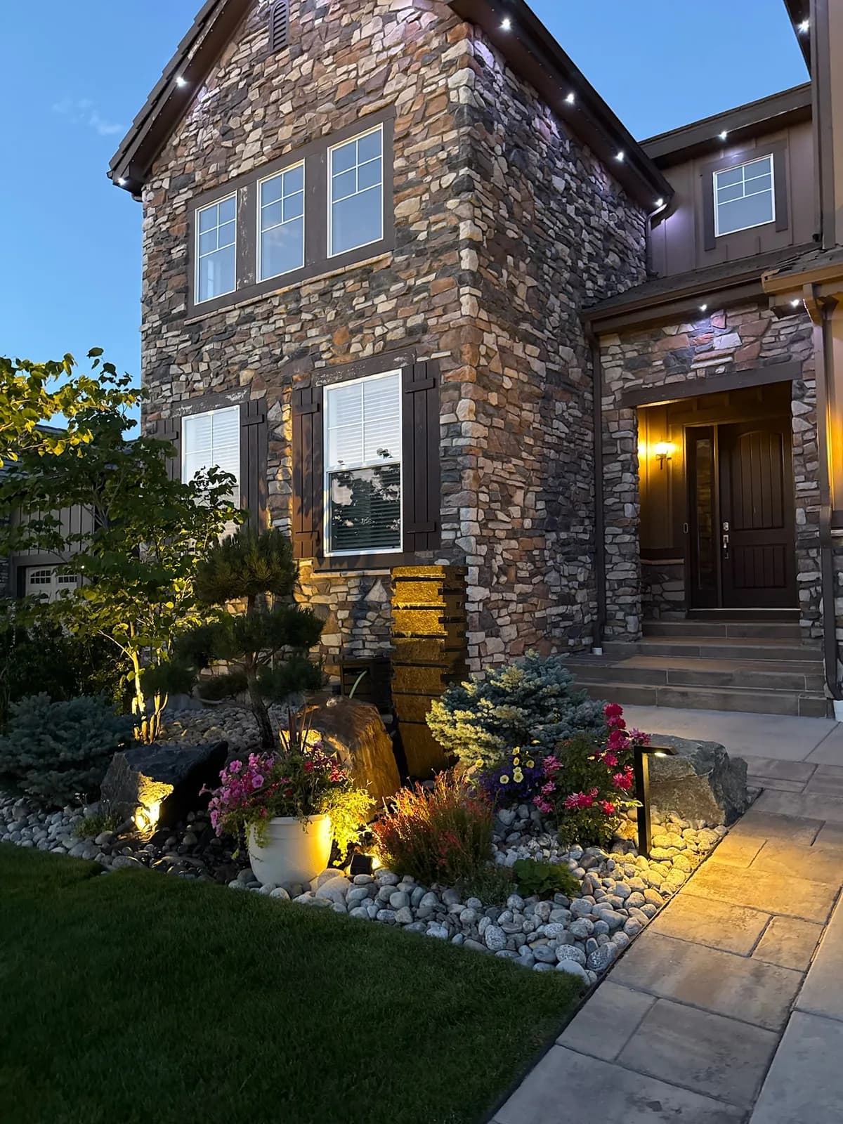 Stone-front home entry at dusk with landscape lighting, a water feature, and layered evergreen and flower plantings