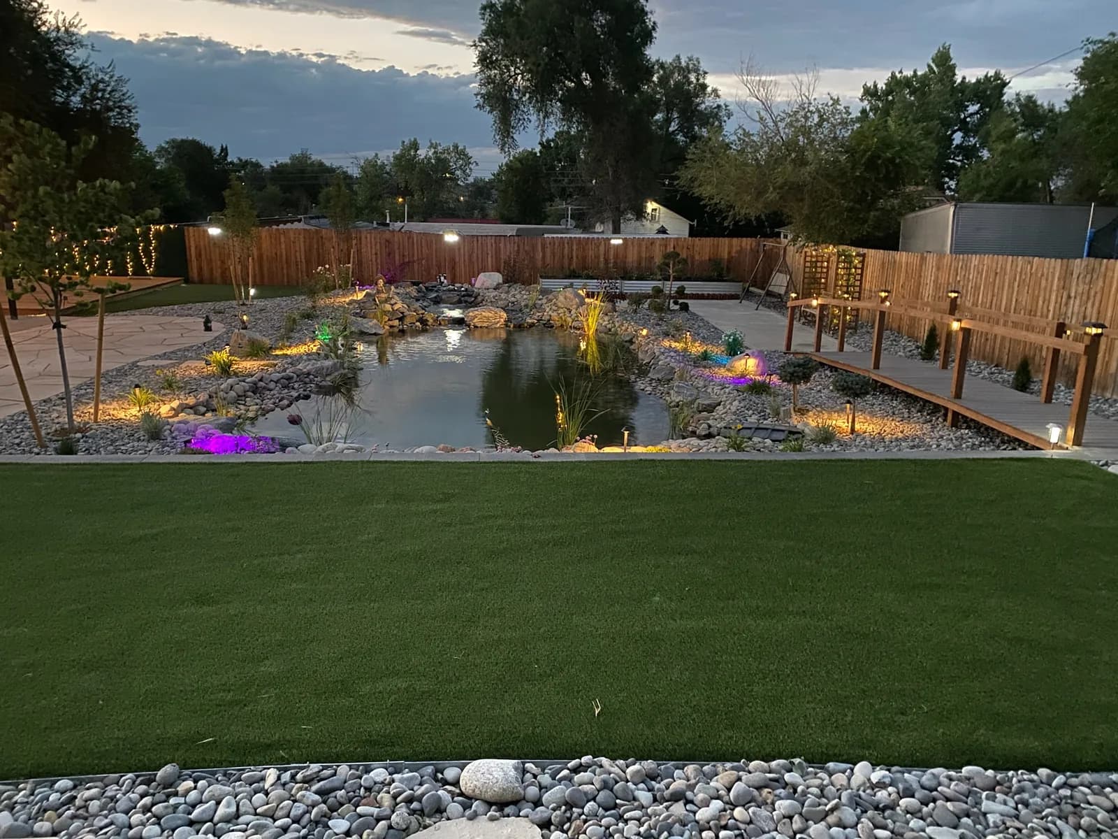 Large landscaped backyard pond lit at dusk, ringed with river rock and lawn